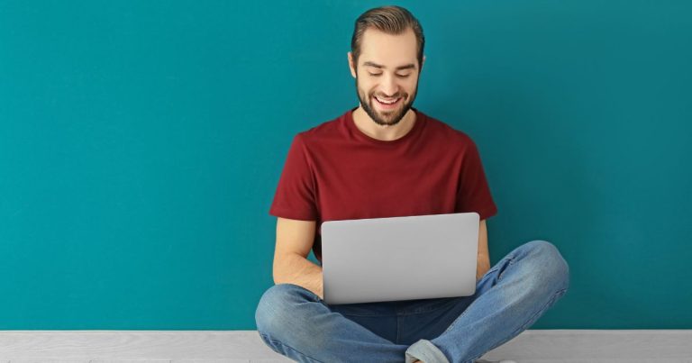 A young man in a red t-shirt and jeans, sitting cross-legged working on a laptop in his lap working on SEO for his website so he can attract more visitors to his website and business.